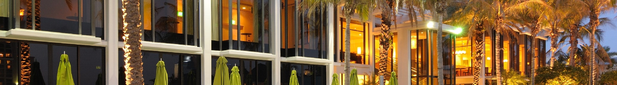 Row of modern buildings with large windows illuminated at dusk, lined by palm trees and green umbrellas on a patio—a perfect spot for professionals to discuss effective tax planning strategies.