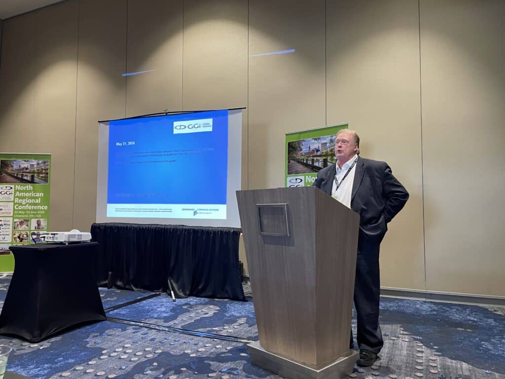 A man in a suit stands at a podium giving a presentation in a conference room, with a projected slide and banners in the background.