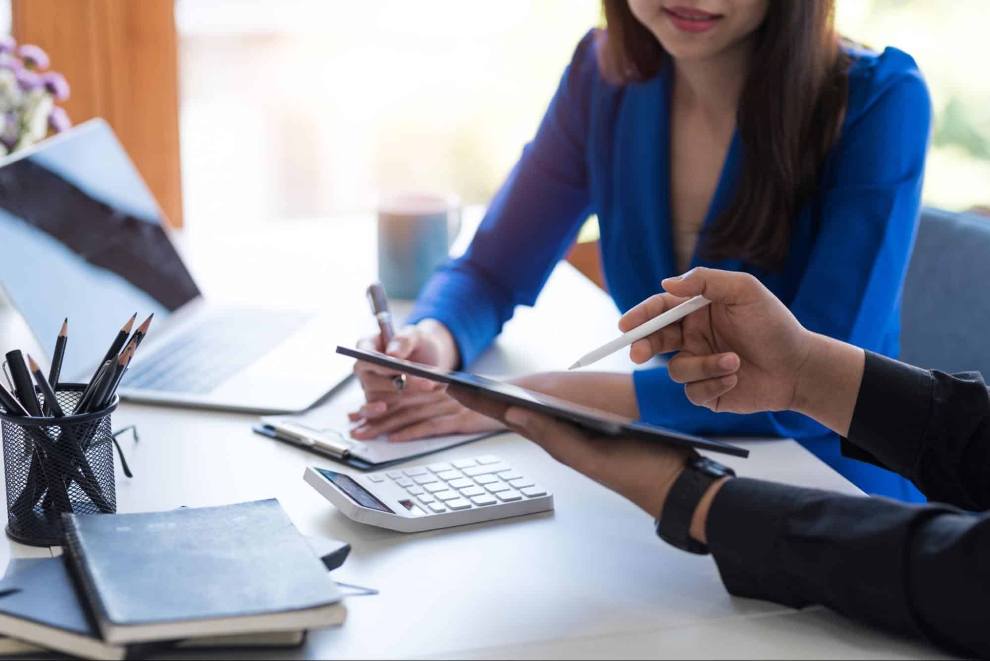 Two people sit at a desk with a laptop, calculator, notebook, and tablet, discussing and reviewing information together as they focus on Forensic Accounting Services.