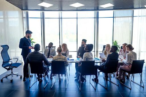 A group of people in business attire sit around a conference table in a modern office while one person stands and speaks to them.