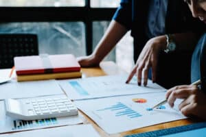 Two people review financial charts and graphs on paper at a desk with a calculator, pen, and closed folders, focusing on year end tax planning.