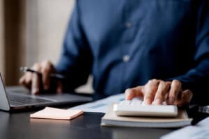 A person researches how to invest in REITs, using a calculator and a laptop at a desk scattered with documents and sticky notes.