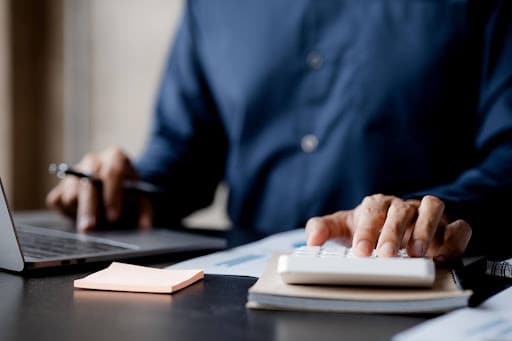 A person researches how to invest in REITs, using a calculator and a laptop at a desk scattered with documents and sticky notes.