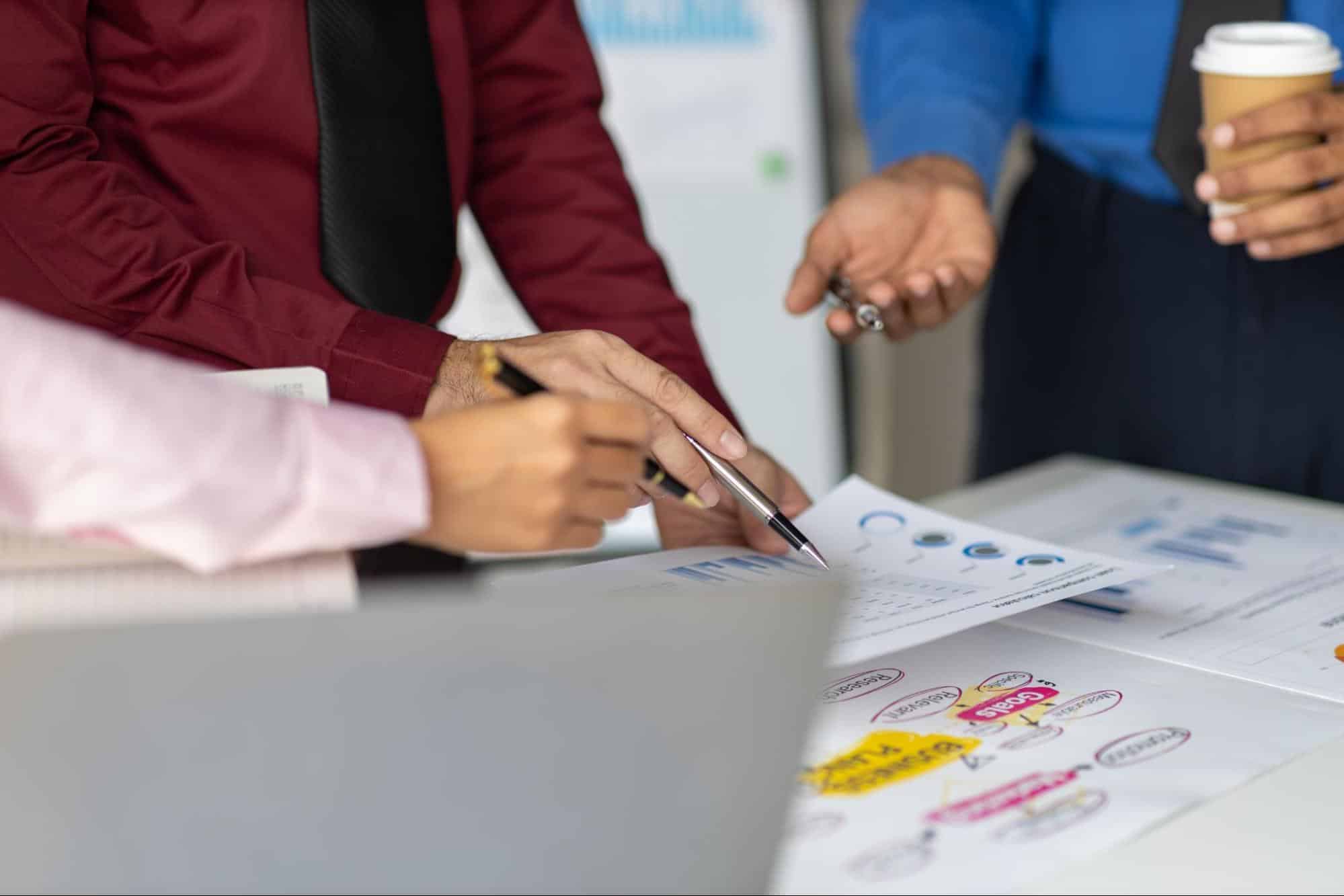 Three people in business attire discuss and point at printed charts and graphs on a desk; one person holds a coffee cup.