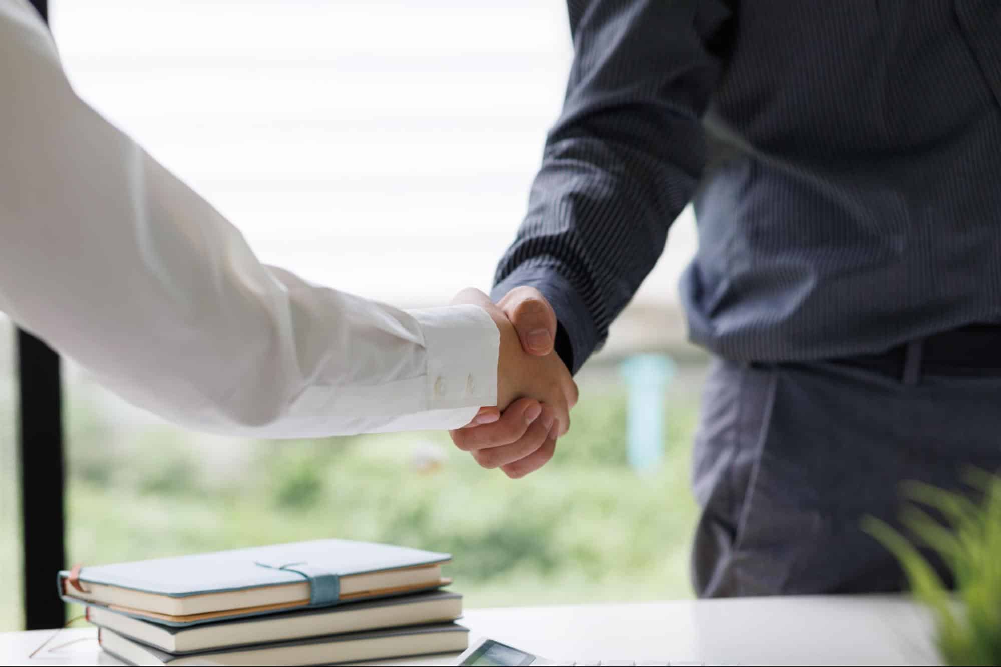 Two people in business attire shake hands across a desk with stacked notebooks in the foreground.