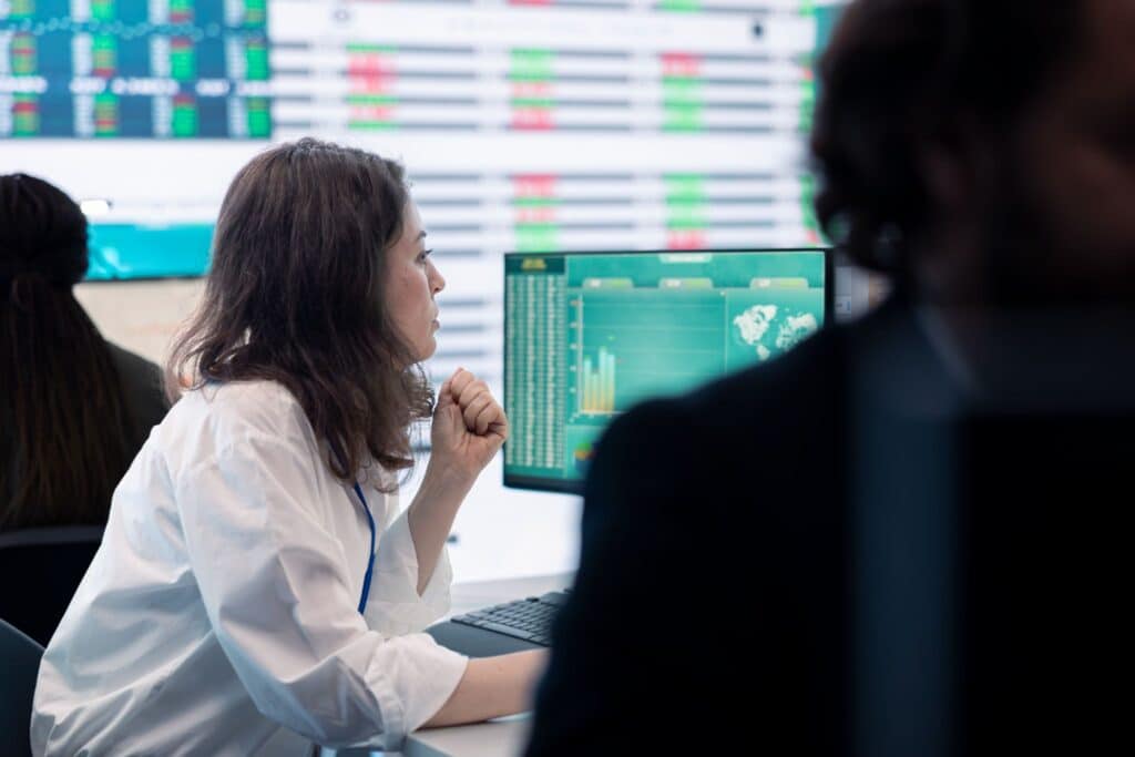 A woman in a white shirt looks at computer screens displaying Financial Irregularities in an office with large digital displays in the background.