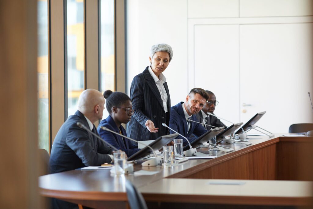 A woman in a suit stands and speaks to four seated colleagues at a conference table with monitors in a modern office setting.