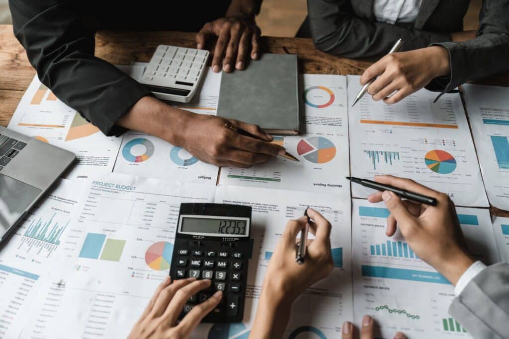 Four people analyze financial charts and graphs on printed papers, using calculators and pens, at a table during a business meeting.