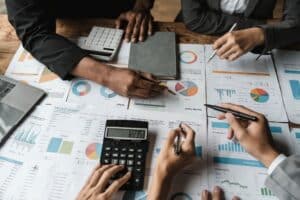 Four people analyze financial charts and graphs on printed papers, using calculators and pens, at a table during a business meeting.